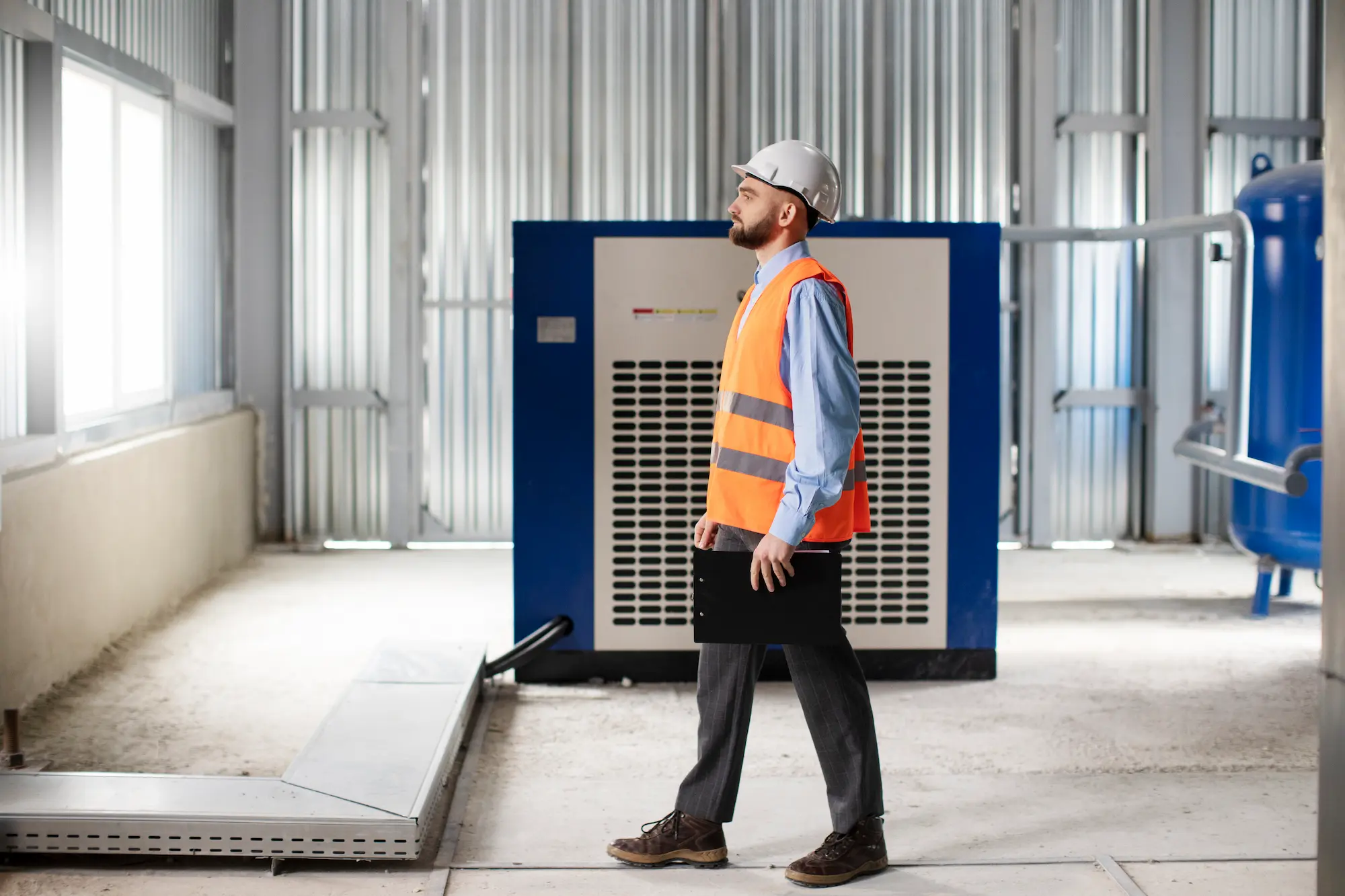 Man in safety vest walking in industrial setting with clipboard.